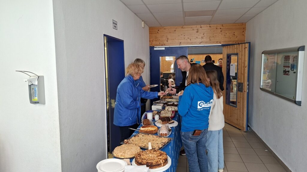 Several people stand at a table covered with cakes and desserts in a hallway. Two people behind the table serve baked goods, while others wait in line. The setting appears to be a community or school event.