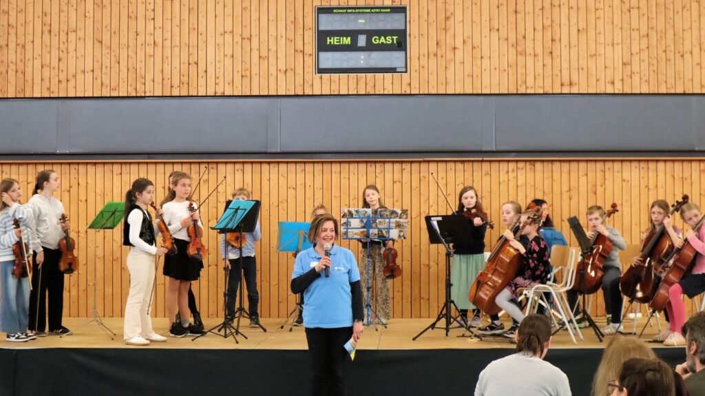 A group of children with string instruments stand on a stage in a gymnasium, while a woman in a blue shirt speaks into a microphone at the front. An electronic scoreboard hangs on the wooden wall behind them.