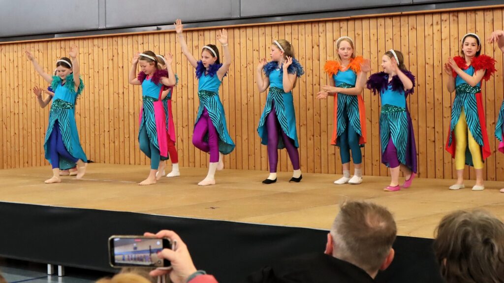 A group of young girls in colorful costumes and feather boas perform a dance on stage, while audience members in the foreground watch and record with a smartphone.