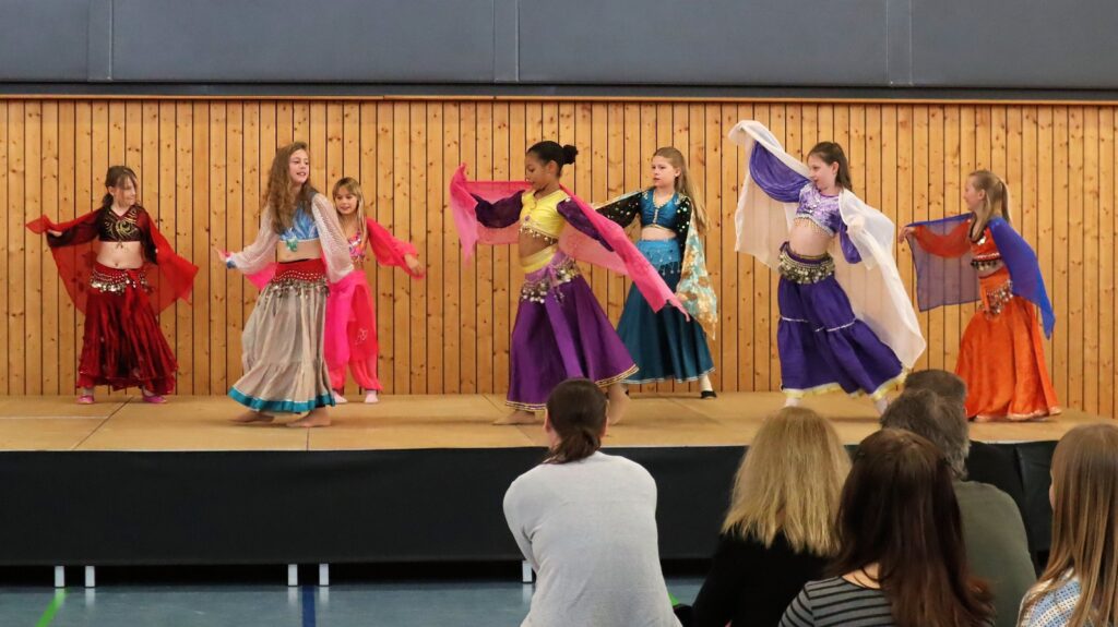 Six young girls in colorful belly dance costumes perform on a stage with wooden paneling, holding sheer scarves, while a small audience watches them from the floor.