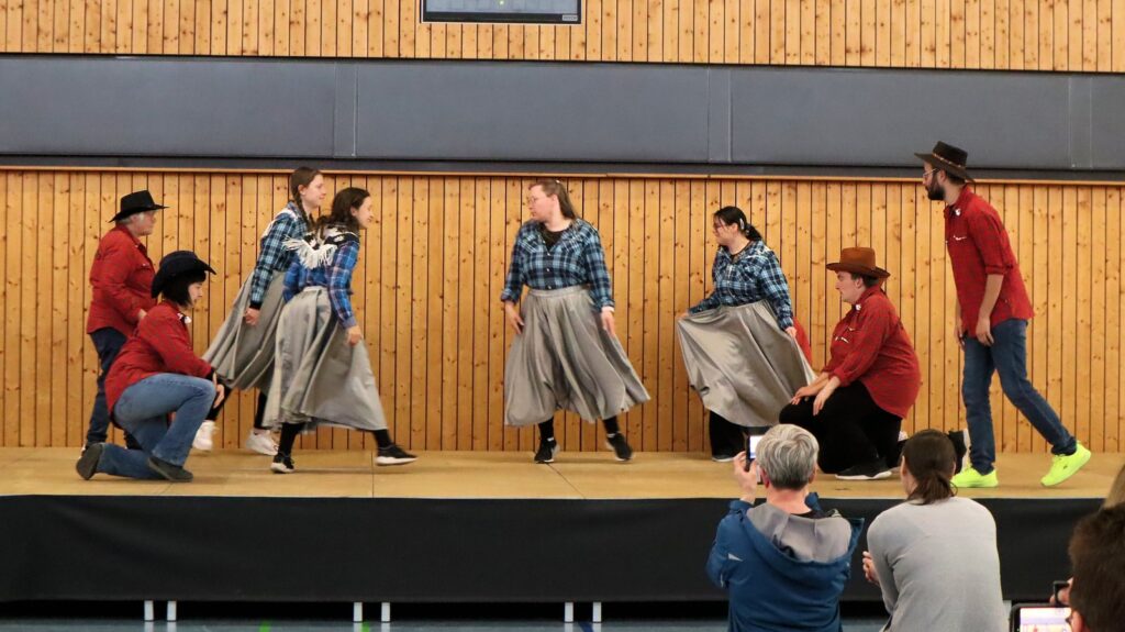 A group of people in western-style clothing perform a dance on a wooden stage, with several women twirling long skirts. Two people in the foreground sit and watch, one holding up a phone to record.