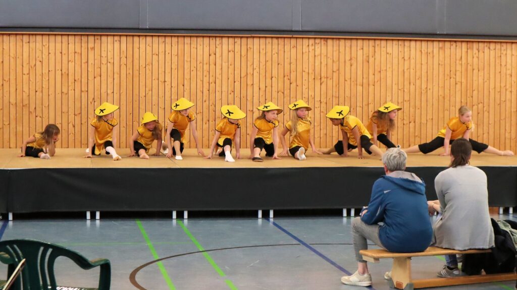 A group of children wearing yellow shirts and hats performs a dance routine on a stage in a gymnasium, with two adults seated on a bench watching them.