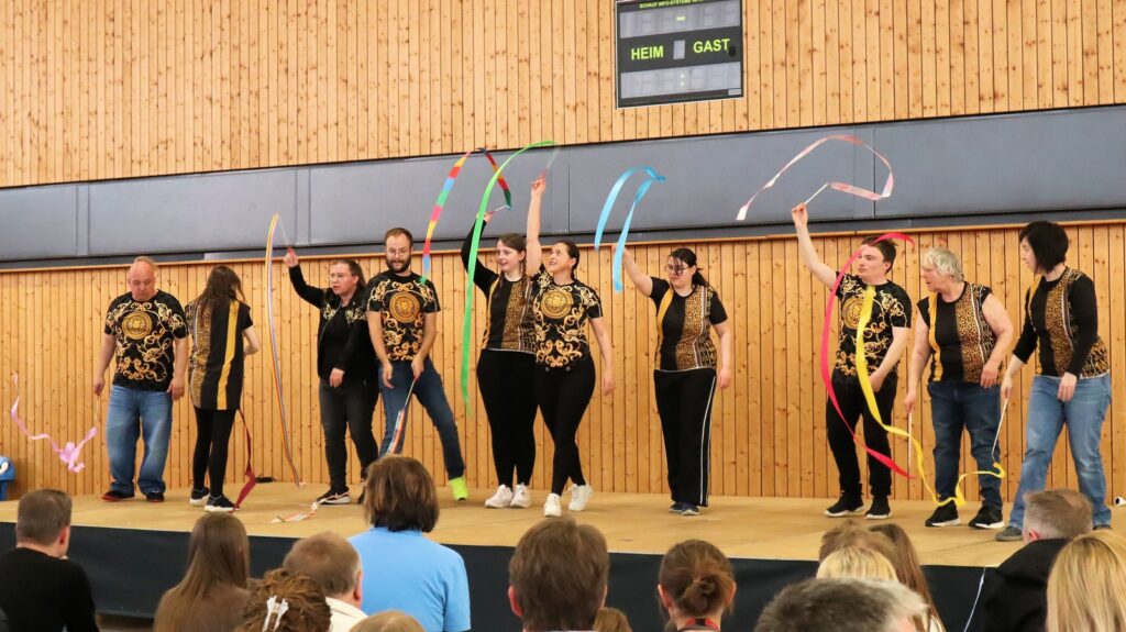 A group of people perform onstage in front of an audience, waving colorful ribbons. They wear matching black and gold shirts. The background is a wooden wall with a scoreboard above.