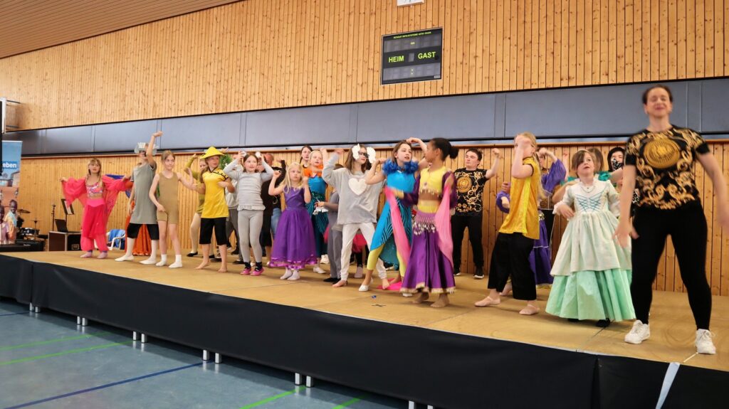 A diverse group of people in colorful costumes perform energetically on a stage in a gymnasium, with a wooden wall and scoreboard in the background.
