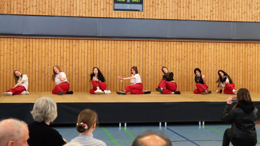 Seven young dancers in matching red pants and white or black tops perform a synchronized routine on a stage, while an audience watches from the foreground. The background features a wooden wall and an electronic scoreboard.