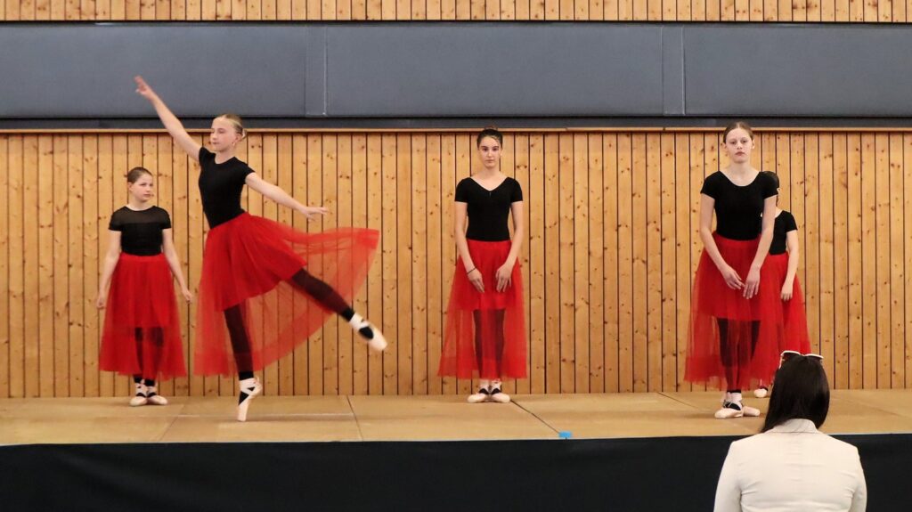 Four young ballet dancers in black tops and red tulle skirts stand on a stage with a wooden wall background; one dancer is performing a leap while the others stand still. A person is seated in front, watching.