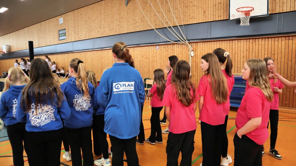 A group of girls stands in a gymnasium, some wearing matching blue jackets and others in pink shirts, facing away from the camera. A basketball hoop and wooden walls are visible in the background.