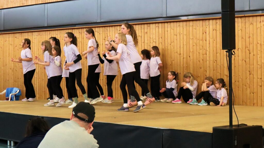 A group of children in matching shirts and leggings perform a dance routine on stage, while several other children sit along the wall, watching or resting. An audience member watches from the foreground.