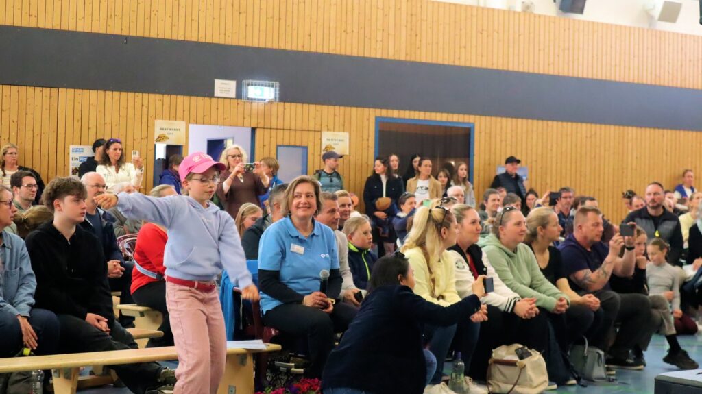 A crowd of people, including children and adults, sits on benches in a gymnasium. A girl in a pink cap and light blue sweater stands in the foreground with her arm raised. Everyone is watching something out of frame.