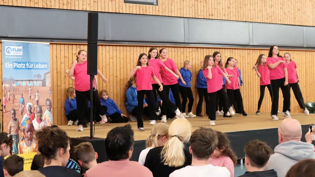 A group of girls in pink shirts dances on a stage, while others in blue shirts kneel behind them. Audience members watch from the foreground. A banner with the words Ein Platz für Leben is visible on the left.