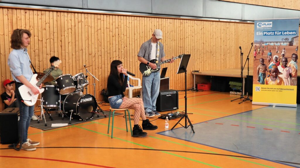 A youth band performs indoors; a girl sings into a microphone, backed by guitar, bass, and drums. A banner from Plan International stands nearby, showing children and the text Ein Platz für Leben.