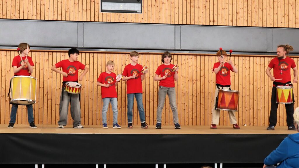 A group of seven people, wearing matching red shirts, stand on a stage playing drums and percussion instruments against a wooden panel backdrop.