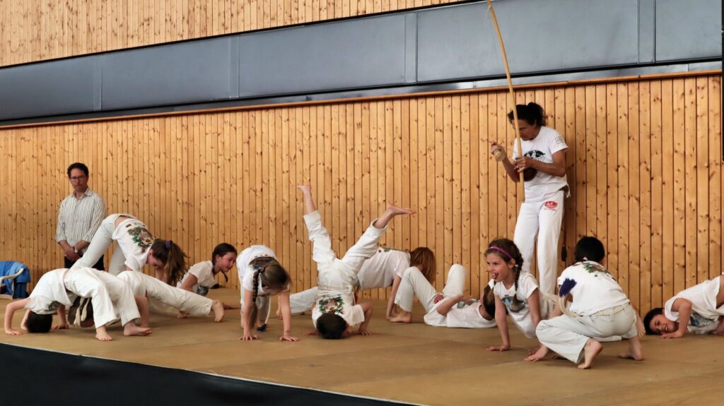 A group of children in white clothing practice capoeira moves on a gym mat, with an adult playing a berimbau and another adult watching in the background. The setting has a wooden wall and bright, indoor lighting.