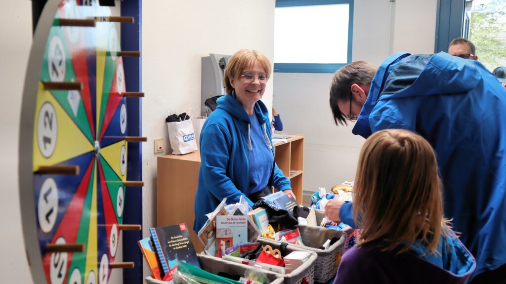 A woman smiles at a table filled with prizes and books while a man and a child look at the items. A colorful prize wheel stands nearby. The scene takes place indoors, near a window.