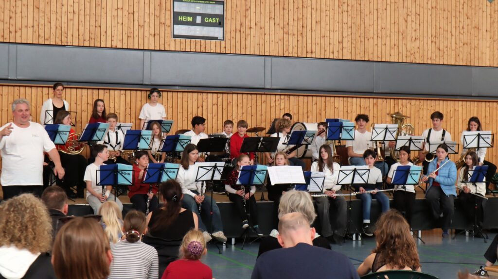 A school band of children performs on stage with various instruments, including clarinets, flutes, and brass, while an adult conductor stands to the left; an audience watches in a gymnasium setting.