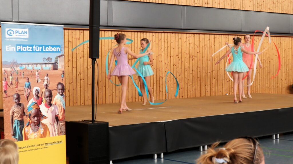 Four young girls in colorful ballet costumes perform a ribbon dance on a stage. To the left, a vertical banner with images of children and German text promotes a charity organization. Audience members watch from below.
