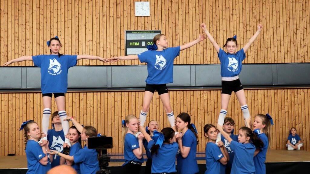 A group of young cheerleaders in blue uniforms performs a stunt indoors; two girls stand balanced on their teammates shoulders, holding hands and raising their arms, with a gymnasium wall in the background.