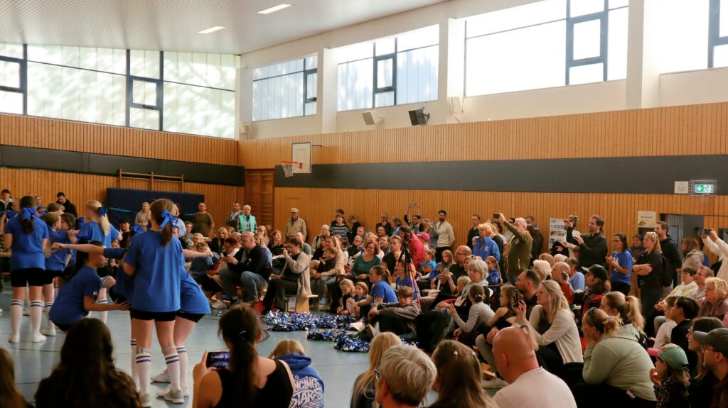 A large group of people sit and stand in a gymnasium, watching cheerleaders in blue uniforms perform. Some spectators are taking photos or videos. The gym has high windows and wooden paneling on the walls.