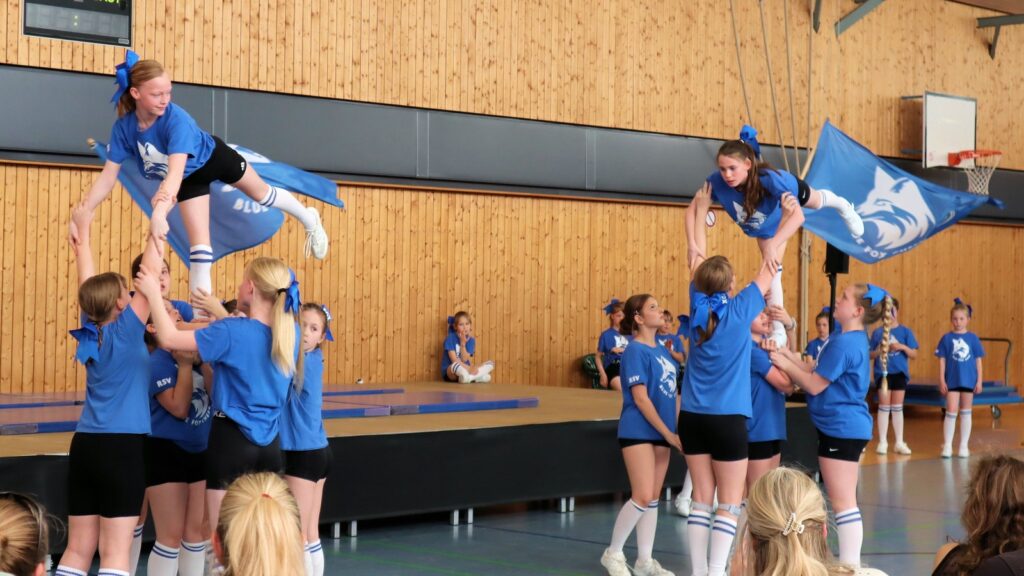 Cheerleaders in blue uniforms perform a routine indoors, lifting teammates into the air. Some hold flags, and a small audience watches. The background features a wooden wall and a stage.